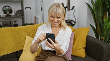 Şık bir iç mekan içinde neşeli bir sarışın kadın akıllı telefonunu kullanırken gülümsüyor, koltukta canlı minderlerle oturuyor..