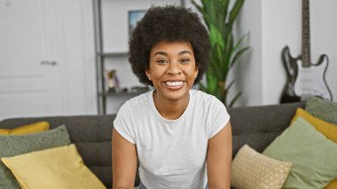 Kıvırcık saçlı, gülümseyen Afro-Amerikan kadın oturma odasında poz veriyor.