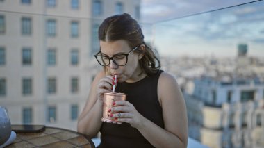 Güzel İspanyol bir kadın, gözlük takıyor, çatıdaki terasta tropik bir kokteylin tadını çıkarıyor, şehrin yukarısında şen şakrak bir kutlama.