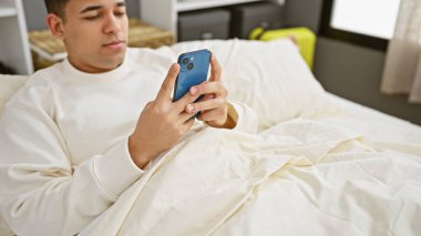 Genç adam beyaz bir yatak odasında dinlenirken akıllı telefon kullanıyor.