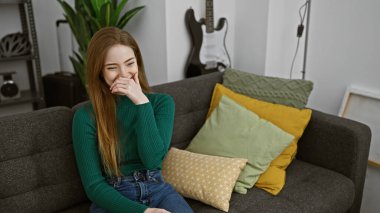 İçerideki gri bir kanepede, renkli yastıklarla çevrili, gülümseyen beyaz bir kadın..