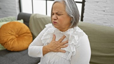 İyi aydınlatılmış bir oturma odasındaki kanepede otururken göğüs ağrısı çeken son sınıf öğrencisi bir kadın..