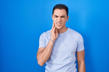 Young hispanic man standing over blue background touching mouth with hand with painful expression because of toothache or dental illness on teeth. dentist 