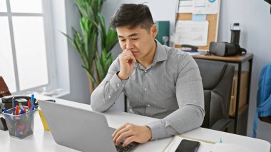 Modern ofis ortamında dizüstü bilgisayarla çalışan düşünceli Asyalı bir adam profesyonelliği ve konsantrasyonu yansıtıyor..