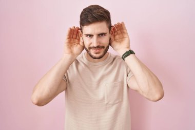 Sakallı İspanyol bir adam pembe arka planda dikilmiş iki elini de kulak hareketi yaparken duymaya çalışıyor, dedikoduya meraklı. İşitme sorunu, sağır 