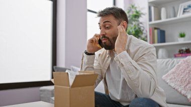 Young hispanic man speaking on the phone unpacking cardboard box at home clipart