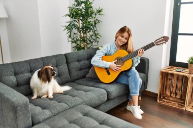 Klasik gitar çalan beyaz bir kadın evdeki kanepede köpeğiyle oturuyor.