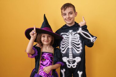 Neşeli erkek ve kız kardeşler, ürkütücü cadılar bayramı kostümleri içinde, güvenle işaret ediyor ve neşeyle parlıyorlar. Güneşli, sarı ve izole bir arkaplan..