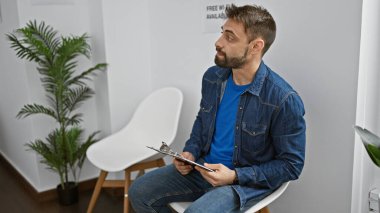 Yakışıklı, İspanyol asıllı, sakallı, ciddi yüzlü bekleme odasında dinlenen bir adam. Sandalyeye otururken dinlenmek, içeride röportaj belgeleriyle pano tutmak..