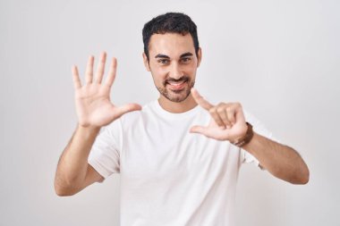 Beyaz arka planda duran yakışıklı İspanyol adam yedinci parmaklarını gösterip işaret ederken kendine güvenen ve mutlu bir şekilde gülümsüyor.. 