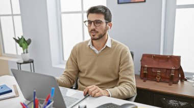 Gözlüklü İspanyol adam modern ofiste dizüstü bilgisayarda çalışıyor, profesyonellik ve odaklanma gösteriyor..