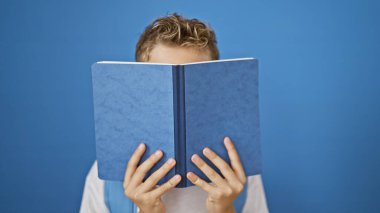 Sevimli sarışın öğrenci, hevesli bir öğrenci, akademik çalışmaya dalmış, zeki bir şekilde küçük yüzünü bir kitapla örterek, parlak mavi arka plan duvarına karşı duruyor..