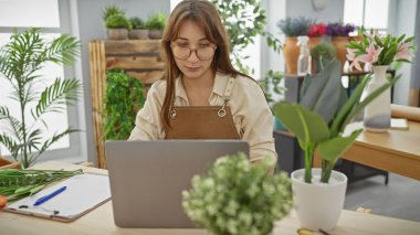 Çiçekçi dükkanında dizüstü bilgisayarda çalışan gözlüklü genç bir kadın. Etrafı çiçek ve bitkilerle çevrili..