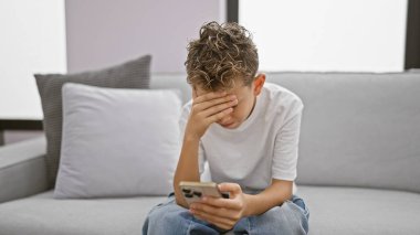Sevimli sarışın çocuk, stresli ve üzgün, akıllı telefon kullanarak evdeki kanepede oturuyor. Teknoloji stresinin ortasında ciddi bir ifade gösteren bir mesaj yazan endişeli bir çocuk..