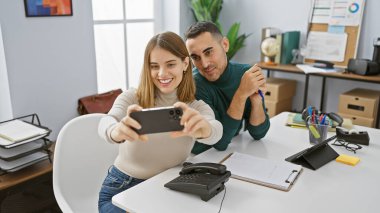 Kadın ve erkek iş arkadaşları, etraflarında teknoloji ve kırtasiye malzemeleriyle bir ofis masasında selfie çekiyorlar..