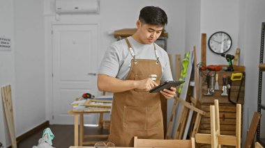 Marangozluk atölyesinde tablet kullanan önlüklü bir Asyalı..
