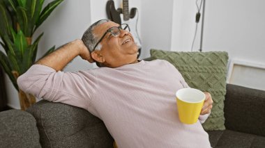 Rahat, orta yaşlı, gri saçlı bir adam oturma odasında mola verip, elinde sarı bir kupayla mutlu mesut gülümsüyor..