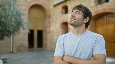 Sarışın sakallı neşeli genç adam, kollarını kavuşturarak gülümsüyor, yeşil ağaçlara bakarak yeşil şehir parkında gelişigüzel dikilirken mutluluğun tadını çıkarıyor..