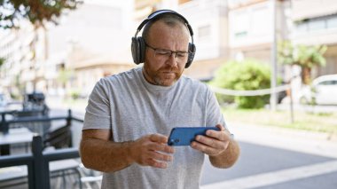 Beyaz adam sokakta akıllı telefon kullanıyor. Kulaklık takıyor.
