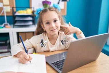 Masada oturmuş dizüstü bilgisayarla ödev yapan küçük bir kız mutlu bir şekilde eli ve parmağı işaret ediyor. 