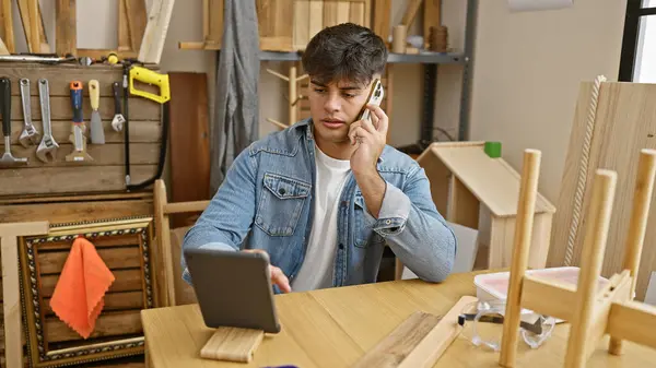 Hardworking young hispanic man, a dedicated builder and carpenter engaged in his profession, talking on smartphone and using touchpad amidst the lumber and timber at his workshop