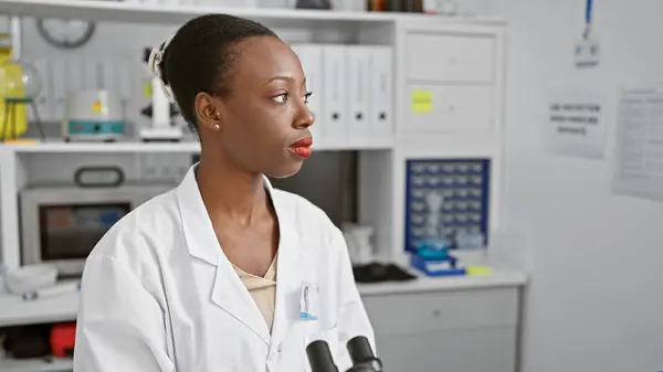 Serious african american woman scientist at work, sitting in a lab engaged in medical research, microscope at hand, making immense strides in biology analysis