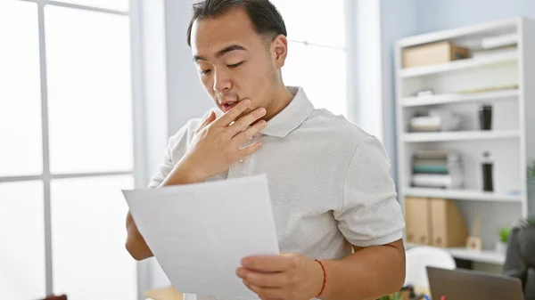 Handsome young chinese man, a focused professional, experiencing a frustrating day at work. upset yet staying concentrated, he\'s reading a worrying document in the indoor office room.