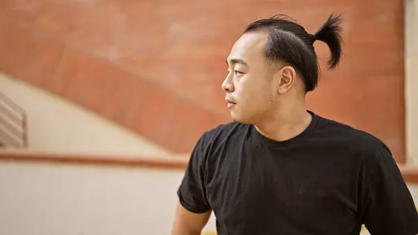 Handsome, cool, young chinese man, with trendy pigtail hairstyle, standing outside on sunny, urban street. serious expression as he concentrates, looking to the side with a hint of mystery.