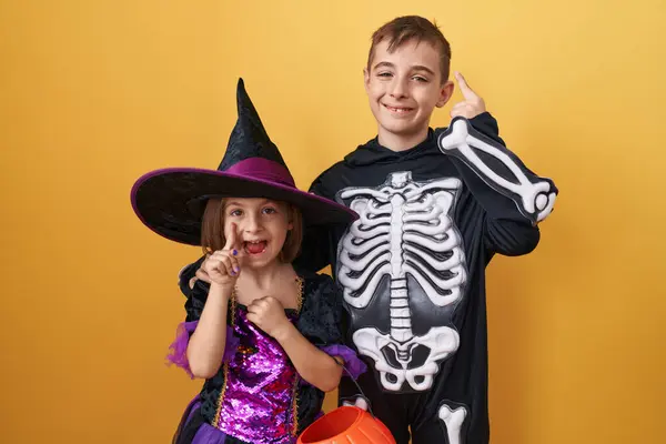 stock image Cheeky brother and sister duo wearing spooky halloween costumes present a silly question with merry faces, finger-pointing at number one on a vibrant, isolated yellow background