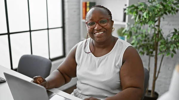 Confident african american woman boss, rocking braids and glasses, smiling as she\'s working online on her laptop at her elegant office desk.