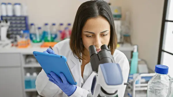 若いヒスパニック系女性科学者が 実験室でタブレットを保持しながら顕微鏡でサンプルを分析 — ストック写真