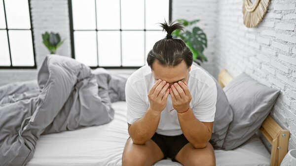 Тревога захватывает молодого китайца, борящегося со стрессовой проблемой, выглядит взволнованным, сидя на кровати в унылой спальне утром, проявляя признаки эмоционального стресса.