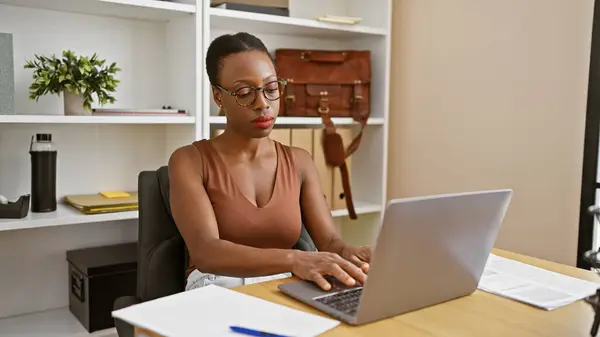 Hardworking african american woman boss, beautiful and serious, working online on laptop at office table with focused face