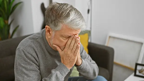 stock image A distressed senior man indoors covering face with hands, showing signs of sadness or headache in a living room.