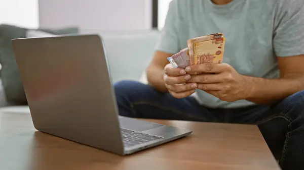 Young man sitting at home, banking online on his laptop, holding wads of rich mexican peso banknotes on his apartment\'s sofa.