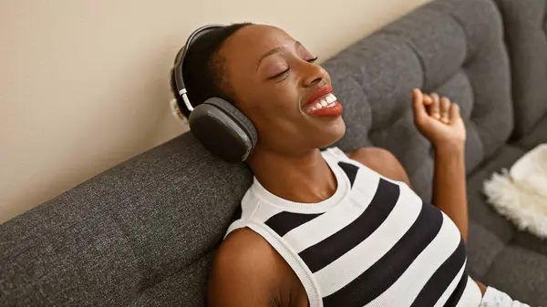 Cheerful african american woman enjoying music, positively vibing, sitting on cozy sofa at home, immersed in sound of joy, listening on cool gadget, adding fun to lifestyle with a confident smile