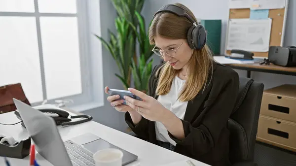 Ofisteki Genç Sarışın Bir Kadın Akıllı Telefonuna Kulaklıklarına Laptopuna Kahvesine — Stok fotoğraf