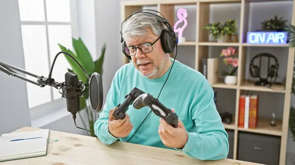 stock image A grey-haired man examines game controllers while recording in an indoor studio with an 'on air' sign.