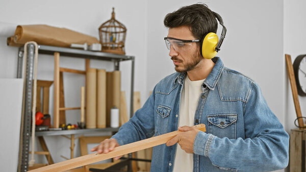 Handsome hispanic man with beard wearing safety glasses and headphones examines wood in a carpentry workshop