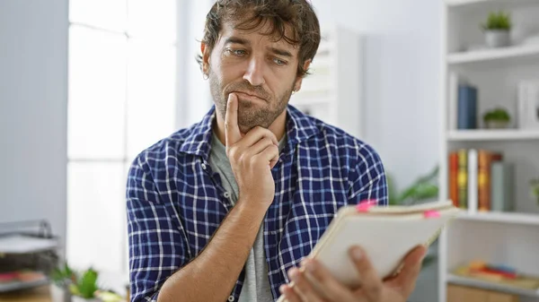Worried, focused young man working hard at his job in the office, reading paperwork and thinking over his notebook, blond beard adding to his professional portrait
