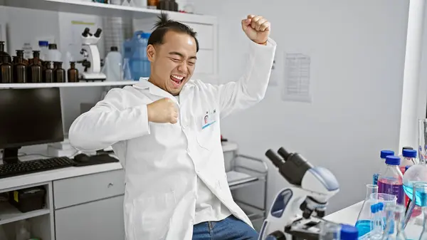Smiling young chinese scientist celebrating major discovery with microscope at lab, embodying the winning spirit of medical research