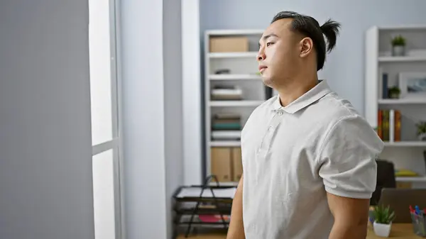 Young, handsome chinese business worker in pigtails looking through the office window with a serious yet relaxed gaze, throwing his successful work side indoors
