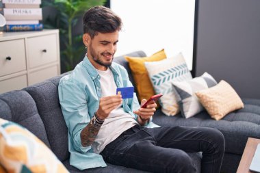 Genç İspanyol adam akıllı telefon ve kredi kartı kullanıyor. Evdeki kanepede oturuyor.