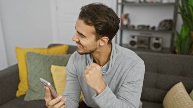 Gülümseyen, sakallı genç bir İspanyol modern bir oturma odasında telefon tutarken iyi haberi kutluyor..