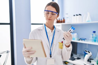 Young caucasian woman scientist holding test tubes reading notebook at laboratory