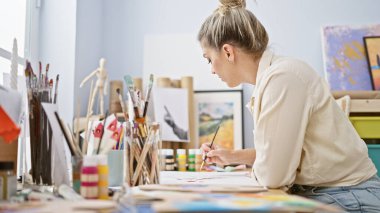 Sanat stüdyosunda genç sarışın kadın ressam ciddi yüzüyle deftere çizim yapıyor.