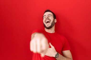 Gündelik kırmızı tişört giyen genç İspanyol adam sana gülüyor, eli vücudunun üzerinde kamerayı işaret ediyor, utanç ifadesi. 