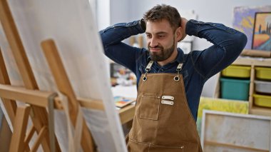 Rahatlamış genç İspanyol bir adam, yakışıklı sakallı bir ressam portresi, resim stüdyosunda fırçalarla resim yapmaya odaklanmış, tuval başyapıtını mükemmelleştiriyor.