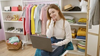 Gülümseyen genç bir kadın, şık kıyafetler ve aksesuarlarla çevrili rahat ve düzenli bir soyunma odasında dizüstü bilgisayarla oturuyor..