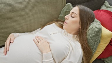 Kanepede dinlenen, renkli yastıklarla kaplanmış, samimi bir ev ortamını tasvir eden, dingin bir İspanyol hamile kadın..